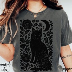 May include: A gray t-shirt featuring a black cat design surrounded by flowers and a crescent moon. The words "comfort colors" and "pepper" are printed on the shirt.