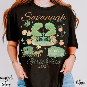 May include: Black t-shirt with a graphic design featuring the text "Savannah Georgia Girls Trip 2025." The design includes a fountain, horse-drawn carriage, trolley, and peach illustrations in shades of green, peach, and teal.