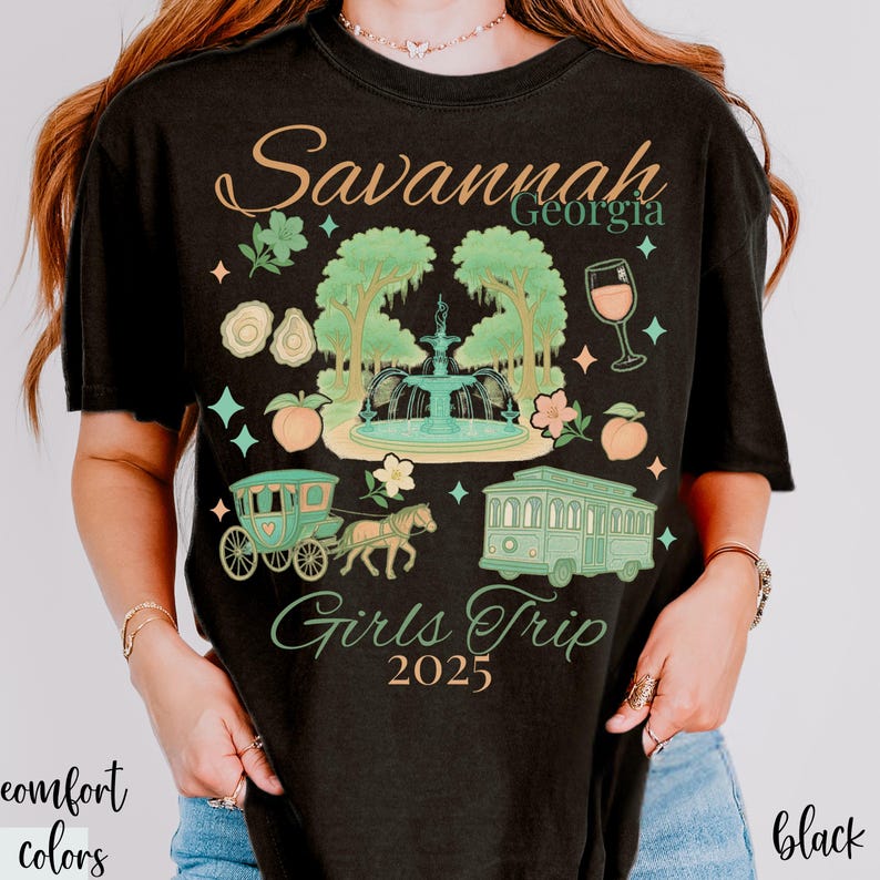 May include: Black t-shirt with a graphic design featuring the text "Savannah Georgia Girls Trip 2025." The design includes a fountain, horse-drawn carriage, trolley, and peach illustrations in shades of green, peach, and teal.