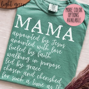 May include: A light green t-shirt with the word "MAMA" in large white letters. Below the word "MAMA" is a list of inspirational words in white script. A pink circle with the text "MORE COLOR OPTIONS AVAILABLE" is in the upper right corner.