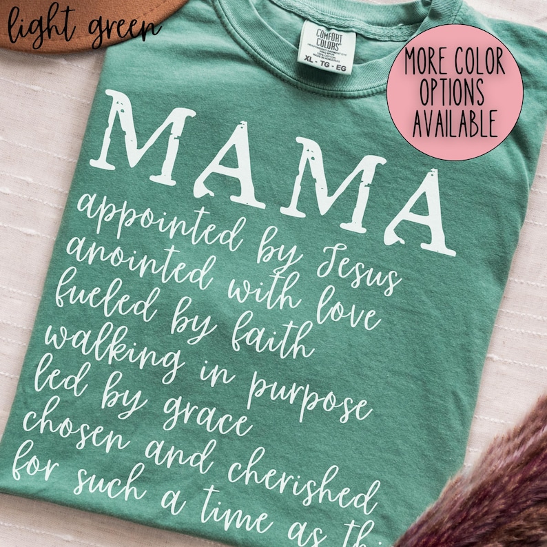 May include: A light green t-shirt with the word "MAMA" in large white letters. Below the word "MAMA" is a list of inspirational words in white script. A pink circle with the text "MORE COLOR OPTIONS AVAILABLE" is in the upper right corner.
