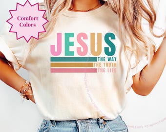 May include: A white t-shirt with the word "JESUS" in colorful letters and the phrase "The Way, The Truth, The Life" in smaller letters below. The shirt is being worn by a person with long blonde hair.
