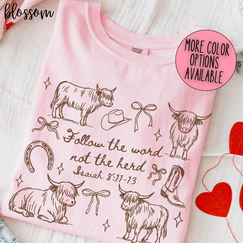 May include: A light pink crew neck t-shirt with a brown graphic design featuring Highland cows, a cowboy hat, a horseshoe, bows, and a cowboy boot. The text "Follow the word not the herd Isaiah 8:11-13" is also printed on the shirt.