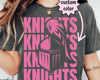 May include: Dark gray t-shirt with the word "KNIGHTS" in pink, along with a pink knight's helmet graphic. The words "comfort colors" and "pepper" are also visible on the shirt.
