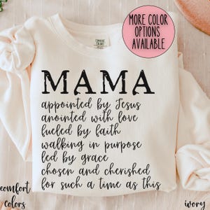 May include: Ivory-colored sweatshirt with the word "MAMA" in distressed black lettering. Below, inspirational text reads "appointed by Jesus, anointed with love, fueled by faith..." A pink circle indicates more color options.