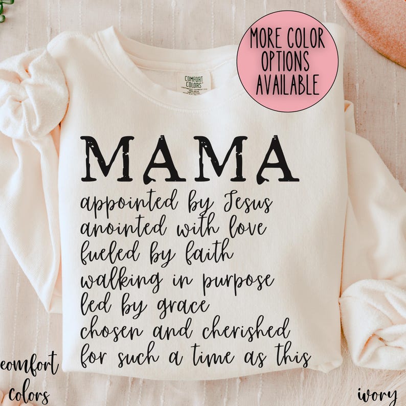 May include: Ivory-colored sweatshirt with the word "MAMA" in distressed black lettering. Below, inspirational text reads "appointed by Jesus, anointed with love, fueled by faith..." A pink circle indicates more color options.