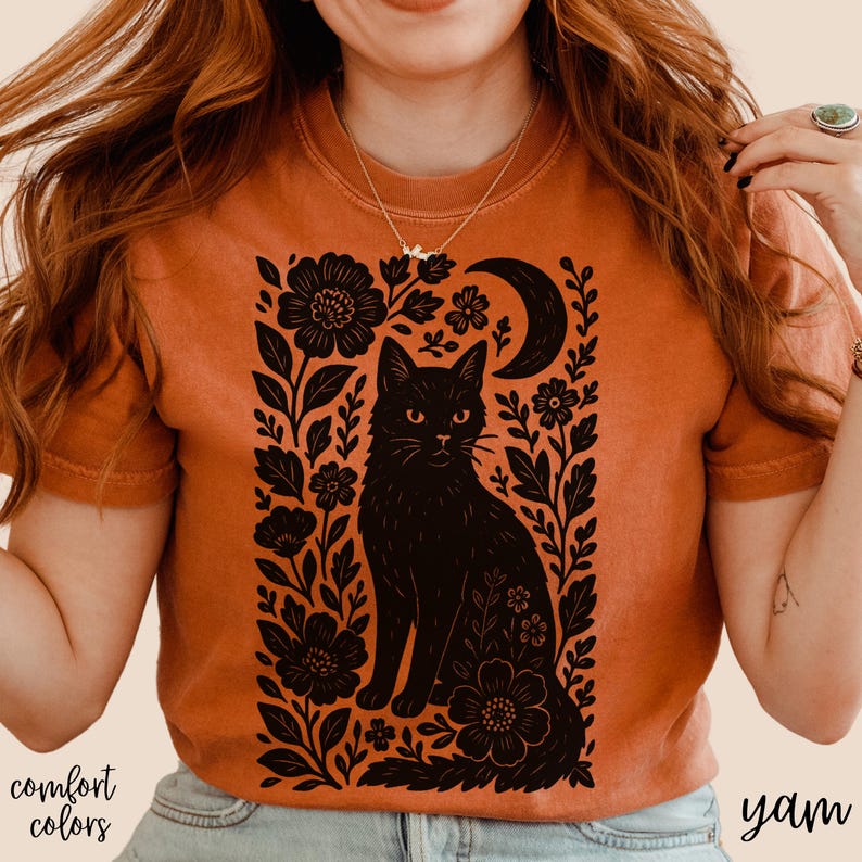 May include: Burnt orange t-shirt featuring a black cat illustration surrounded by floral and crescent moon designs. The words "comfort colors" and "yam" are printed on the shirt. The shirt has short sleeves.