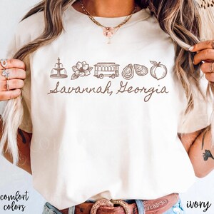 Puede incluir: Camiseta de color marfil con un gr&aacute;fico marr&oacute;n de los monumentos de Savannah, Georgia, incluyendo una fuente, una flor, un tranv&iacute;a, ostras y un melocot&oacute;n. El texto "Savannah, Georgia" est&aacute; escrito debajo. Las palabras "comfort colors" est&aacute;n impresas abajo a la izquierda.