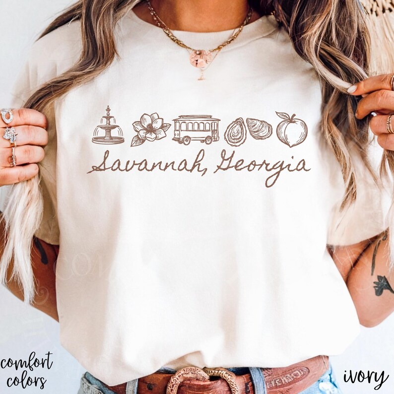 Puede incluir: Camiseta de color marfil con un gr&aacute;fico marr&oacute;n de los monumentos de Savannah, Georgia, incluyendo una fuente, una flor, un tranv&iacute;a, ostras y un melocot&oacute;n. El texto "Savannah, Georgia" est&aacute; escrito debajo. Las palabras "comfort colors" est&aacute;n impresas abajo a la izquierda.