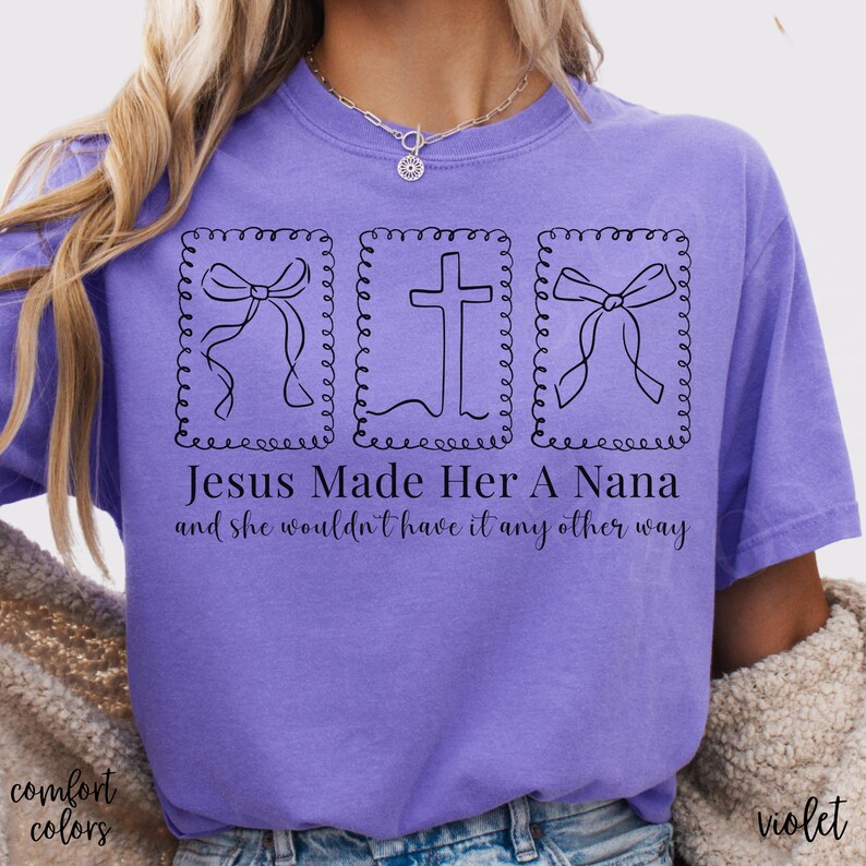 Pu&ograve; includere: Una maglietta viola con la scritta "Jesus Made Her A Nana and she wouldn't have it any other way." La maglietta presenta tre disegni a tratto nero: due fiocchi e una croce, ciascuno all'interno di una cornice decorativa. Sono visibili anche le parole "comfort colors" e "violet".