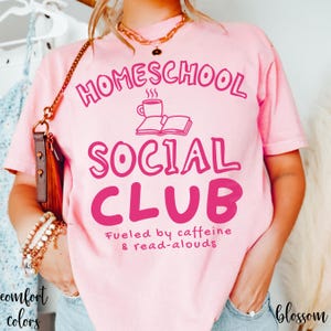 Op de afbeelding: Een lichtroze T-shirt met korte mouwen met de donkerroze tekst HOMESCHOOL SOCIAL CLUB, een afbeelding van een koffiemok en een open boek, en de tekst Fueled by caffeine & read-alouds. Het T-shirt heeft een ronde hals en een ruime pasvorm.