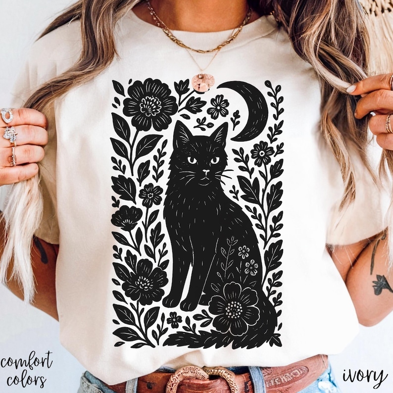 May include: Ivory-colored t-shirt featuring a black cat illustration surrounded by flowers and a crescent moon. The words "comfort colors" are printed on the bottom left. The word "ivory" is printed on the bottom right.