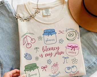 Peut inclure: Sweat-shirt ivoire avec l'inscription "literacy is my jam" et divers graphiques sur le thème de la littératie, notamment des bocaux, des fruits et des fleurs. Le t-shirt comporte également les mots "comprehension", "phonics", "phonemic awareness", "writing", "fluency", "vocabulary", et "language".