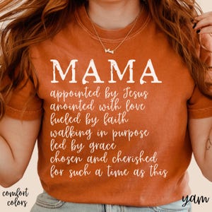May include: An orange-colored t-shirt with the word "MAMA" in large white letters. Below, a list of inspirational phrases is printed in a script font. The words "comfort colors" are printed in the lower left corner.