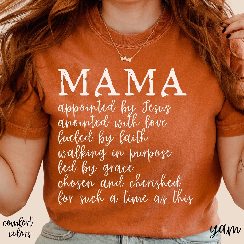 May include: An orange-colored t-shirt with the word "MAMA" in large white letters. Below, a list of inspirational phrases is printed in a script font. The words "comfort colors" are printed in the lower left corner.