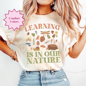 May include: A cream-colored t-shirt with a green graphic that says "LEARNING IS IN OUR NATURE". The graphic is surrounded by small illustrations of nature elements like leaves, mushrooms, and insects.
