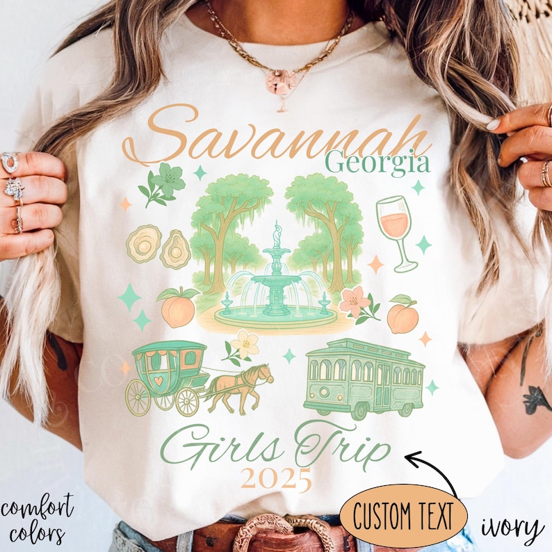 May include: Ivory-colored t-shirt with a graphic design featuring the text "Savannah Georgia Girls Trip 2025." The design includes illustrations of a fountain, trees, a carriage, a trolley, and peaches in shades of green, peach, and teal.