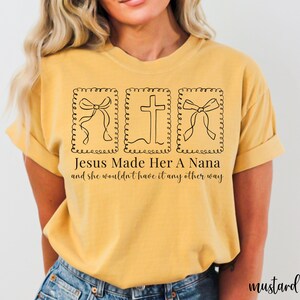 Pu&ograve; includere: T-shirt color senape con un design grafico nero. Il design include due illustrazioni di fiocchi e una croce, incorniciate da un bordo decorativo. Il testo recita: "Jesus Made Her A Nana and she wouldn't have it any other way."