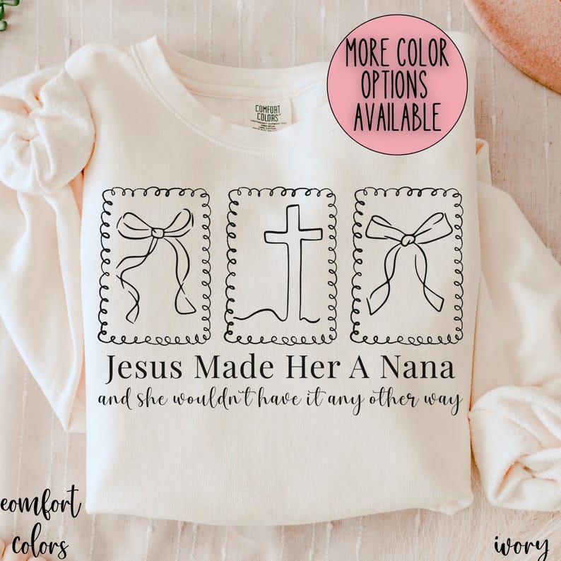 Pu&ograve; includere: Felpa color avorio con la scritta "Jesus Made Her A Nana and she wouldn't have it any other way." Il design include tre illustrazioni incorniciate: due fiocchi e una croce. Un cerchio rosa indica "More Color Options Available."