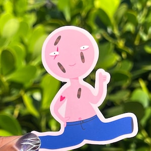 May include: A pink sticker of a cartoon character with a heart tattoo, blue jeans, and a thumbs-up gesture. The character has brown marks and a scar. The sticker is held against a background of green leaves.