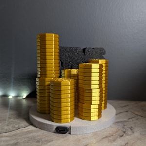 May include: A collection of gold-colored, hexagonal-shaped objects of varying heights, resembling stacks of coins, are arranged on a circular, light gray base. A black, textured block sits behind the gold objects. A USB port is visible on the base.
