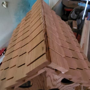 May include: A wooden birdhouse with a shingled roof. The roof is made of individual pieces of wood that are arranged in a pattern. The birdhouse is painted a light brown color.