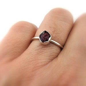 May include: A silver ring with a dark purple gemstone. The ring is on a person's finger.