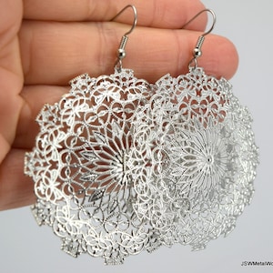 May include: A pair of silver-colored filigree earrings. Each earring features an intricate, openwork design with a circular shape. The earrings have a floral and geometric pattern, with a fish hook style ear wire.