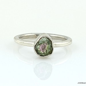 Puede incluir: Un anillo de plata con una piedra preciosa verde y rosa. La piedra preciosa tiene una forma rugosa y natural.