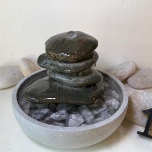 May include: A small, indoor water fountain with a stack of dark gray stones. Water trickles down into a gray stone bowl filled with smaller gray stones. Several smooth, light-colored stones surround the fountain. A black number 1 is next to the fountain.