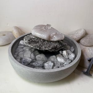 May include: A small, gray concrete bowl filled with water and small stones. Larger, smooth stones surround the bowl. A dark gray metal number 1 is placed near the bowl. The bowl has a natural, zen-like aesthetic.