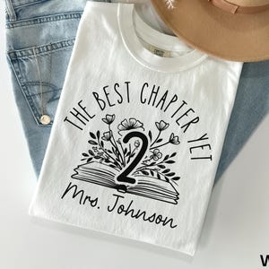 May include: White t-shirt with the text "THE BEST CHAPTER YET 2 Mrs. Johnson" in black. The design includes an open book with floral accents. The shirt is paired with blue jeans and a tan hat.