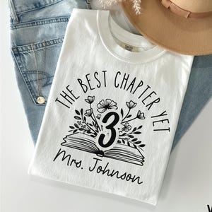 May include: A white t-shirt with the text "THE BEST CHAPTER YET 3 Mrs. Johnson" in black. The design includes an open book, flowers, and the number 3. The shirt is paired with blue jeans and a tan hat.