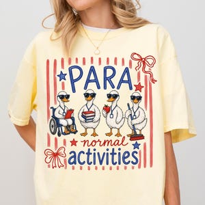 Puede incluir: Camiseta amarilla clara con un gráfico que presenta cuatro patos de dibujos animados con gafas de sol, cada uno realizando una actividad diferente. El texto "PARA normal activities" está impreso en azul y rojo.