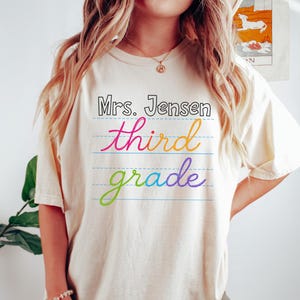 May include: Ivory-colored t-shirt with the text "Mrs. Jensen third grade" in a colorful, handwritten style. The text is on a lined background, giving the impression of a notebook page. The shirt is a casual, oversized fit.