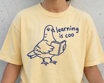 May include: A pale yellow short-sleeve t-shirt featuring a dark blue line art graphic of a pigeon holding an open book. The text "learning is coo" is printed in dark blue next to the pigeon. The shirt has a crew neckline and a relaxed fit.