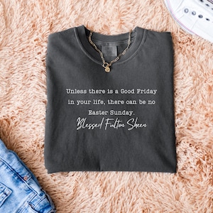 May include: Charcoal grey t-shirt featuring the quote "Unless there is a Good Friday in your life, there can be no Easter Sunday. Blessed Fulton Sheen." A gold necklace is on the shirt. White trainers and blue jeans are also visible.