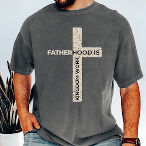 May include: Pepper-colored t-shirt with a white cross graphic. The cross design features the text "FATHERHOOD IS" horizontally and "KINGDOM WORK" vertically. The shirt is a crew neck and has short sleeves.