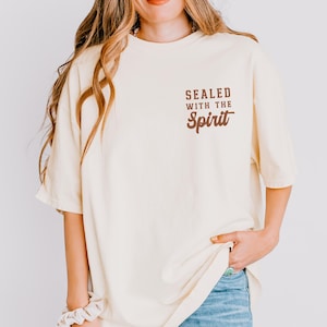 May include: Ivory-colored t-shirt with the text "SEALED WITH THE Spirit" in brown lettering. The shirt has short sleeves and a relaxed fit. The model is wearing blue jeans.