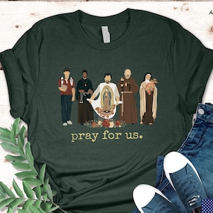 Forest green shirt featuring 5 saints in a row: St. Carlo Acutis, St. Josephine Bakhita, St. Juan Diego, St. Padre Pio and St. Therese of Liseux. The text says "pray for us."