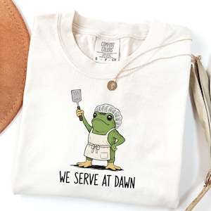 Può includere: Una t-shirt color avorio con la grafica di una rana verde che indossa un cappello da chef e un grembiule bianchi, mentre tiene in mano una spatola. Sotto la rana, il testo recita "WE SERVE AT DAWN". La maglietta ha un'etichetta Comfort Colors.