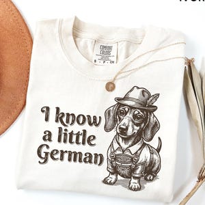 Puede incluir: Una camiseta de color marfil con un gr&aacute;fico marr&oacute;n de un perro salchicha que lleva un sombrero y pantalones de cuero. El texto "I know a little German" est&aacute; impreso en letras marrones a la izquierda del perro. La camiseta tiene una etiqueta.