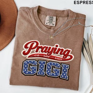 Puede incluir: Camiseta color espresso con las palabras "Praying GIGI" en un diseño rojo y azul. La camiseta tiene cuello redondo y una pequeña etiqueta en la parte superior izquierda. Un collar plateado con un pequeño colgante está sobre la camiseta. Un sombrero marrón está en la esquina superior izquierda.