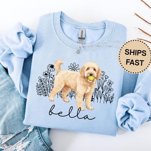 Może przedstawiać: Jasnoniebieska bluza z ilustracją psa golden doodle trzymającego piłkę tenisową, otoczona kwiatami. Imię psa, "bella", jest napisane kursywą. Tekst "SHIPS FAST" znajduje się na naklejce.