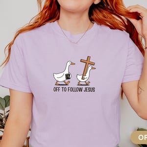 Może przedstawiać: Fioletowa koszulka z zabawnym wzorem dw&oacute;ch białych gęsi. Jedna gęś niesie Biblię, druga drewniany krzyż. Tekst "OFF TO FOLLOW JESUS" jest wydrukowany poniżej. Koszulka jest w kolorze jasnofioletowym.