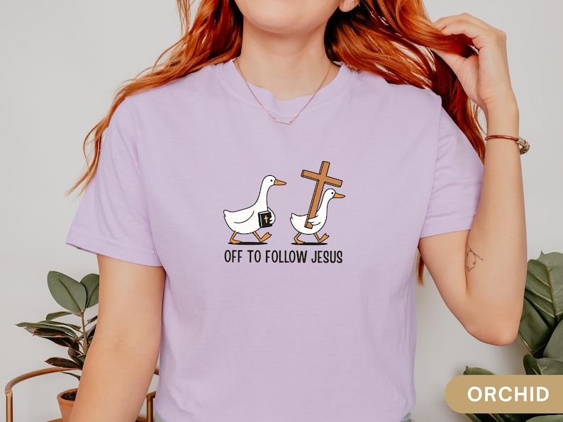 Może przedstawiać: Fioletowa koszulka z zabawnym wzorem dw&oacute;ch białych gęsi. Jedna gęś niesie Biblię, druga drewniany krzyż. Tekst "OFF TO FOLLOW JESUS" jest wydrukowany poniżej. Koszulka jest w kolorze jasnofioletowym.
