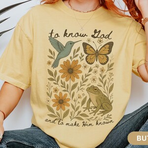 May include: Butter-colored t-shirt featuring a nature-themed graphic with a hummingbird, butterfly, frog, and flowers. The text "to know God and to make Him known" is printed on the shirt.