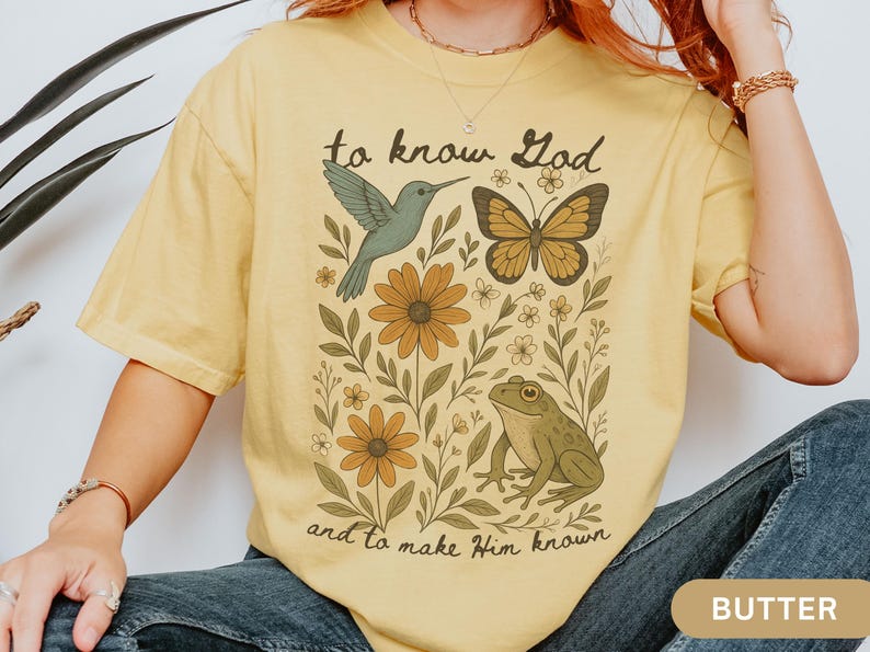 May include: Butter-colored t-shirt featuring a nature-themed graphic with a hummingbird, butterfly, frog, and flowers. The text "to know God and to make Him known" is printed on the shirt.