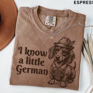 Puede incluir: Una camiseta color espresso con un gr&aacute;fico de un perro salchicha que lleva un sombrero y pantalones de cuero. El texto "I know a little German" est&aacute; impreso en letras de color marr&oacute;n oscuro junto al perro. La camiseta tiene cuello redondo.