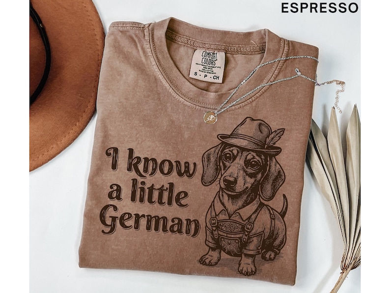 Puede incluir: Una camiseta color espresso con un gr&aacute;fico de un perro salchicha que lleva un sombrero y pantalones de cuero. El texto "I know a little German" est&aacute; impreso en letras de color marr&oacute;n oscuro junto al perro. La camiseta tiene cuello redondo.